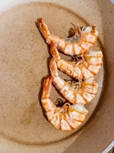 Three cooked prawns are arranged in a circular pattern on a flat, beige dish. The prawns have a reddish-orange hue indicative of being cooked, and their tails are curled.