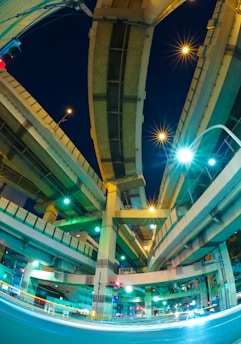 A night scene featuring a complex network of elevated highways intersecting at various angles, illuminated by blue and green streetlights. The perspective is from below, capturing the towering and intricate structure against a dark sky. Light trails from vehicles create dynamic lines at ground level, adding a sense of motion.