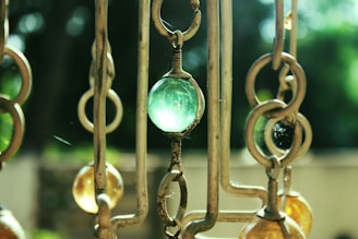 The image depicts a close-up view of a decorative hanging piece made of metal rings and colored glass beads. The central bead is a translucent green, while others are shades of amber. The background is blurred with a mix of dark and light greens, suggesting foliage.