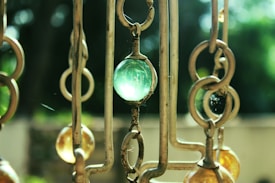 The image depicts a close-up view of a decorative hanging piece made of metal rings and colored glass beads. The central bead is a translucent green, while others are shades of amber. The background is blurred with a mix of dark and light greens, suggesting foliage.
