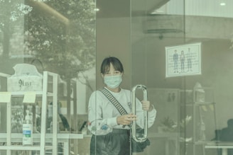 A person wearing a face mask is holding the handle of a glass door. Inside, there is a shelf with items and a bottle of hand sanitizer visible. The interior appears to be a public establishment with signs on the door instructing the use of face masks.