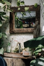 A rustic wooden-framed mirror is surrounded by lush green leaves and vines against a textured wall with some moss. Below the mirror, there's a shelf with a faucet, a bottle of soap, and a small plant. The setting is natural and has a slightly weathered, lived-in appearance.