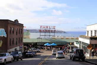 A bustling outdoor market scene with a large, iconic sign displaying 'Public Market' in red letters. The market overlooks a serene body of water with land visible in the background. There are several cars and people walking along the street leading to the market. Buildings on either side frame the scene, featuring a mix of brick and white structures. An American flag can be seen in the distance.