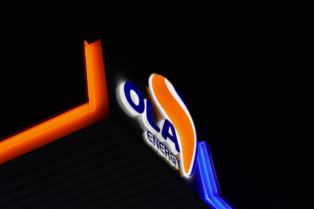 Illuminated signage displaying the word 'OLA' in large blue letters followed by the word 'ENERGY' in smaller letters, with an orange flame-like design curving over the letters. The signage is bordered by bright orange and blue neon lights against a dark background.