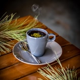 A steaming cup of dark hot beverage garnished with green herbs sits on a wooden table. The drink is in a gray ceramic cup placed on a matching saucer accompanied by a small spoon. Pine-like branches are placed decoratively around the cup.
