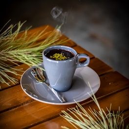 A steaming cup of dark hot beverage garnished with green herbs sits on a wooden table. The drink is in a gray ceramic cup placed on a matching saucer accompanied by a small spoon. Pine-like branches are placed decoratively around the cup.