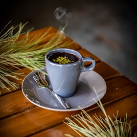 A steaming cup of dark hot beverage garnished with green herbs sits on a wooden table. The drink is in a gray ceramic cup placed on a matching saucer accompanied by a small spoon. Pine-like branches are placed decoratively around the cup.