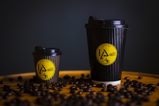 Two black coffee cups, one large and one small, are placed on a surface scattered with coffee beans. The cups have a ribbed texture and feature a yellow circular logo with stylized text and a coffee cup illustration.