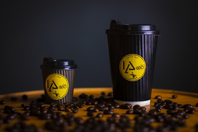 Two black coffee cups, one large and one small, are placed on a surface scattered with coffee beans. The cups have a ribbed texture and feature a yellow circular logo with stylized text and a coffee cup illustration.