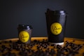Two black coffee cups, one large and one small, are placed on a surface scattered with coffee beans. The cups have a ribbed texture and feature a yellow circular logo with stylized text and a coffee cup illustration.