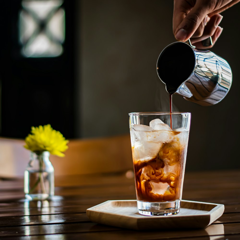 person pouring brown liquid on clear drinking glass