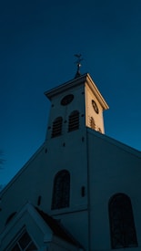 A church building with a tall steeple is illuminated by the soft glow of sunlight, highlighting its architectural features against a clear, deep blue sky. The windows have ornate designs, and the steeple is topped with a distinct weathervane.