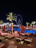 A nighttime scene of a luxurious outdoor pool area with orange umbrellas and loungers, illuminated by palm trees and accent lights. In the background, a large Ferris wheel glows with blue and white lights, enhancing the vibrant cityscape. The setting suggests a relaxing and upscale atmosphere.
