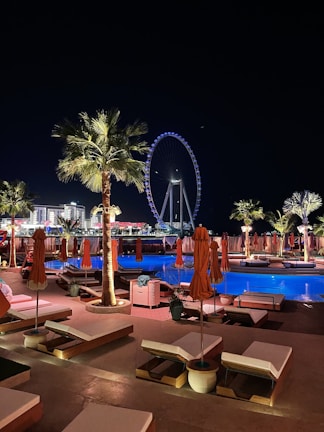 A nighttime scene of a luxurious outdoor pool area with orange umbrellas and loungers, illuminated by palm trees and accent lights. In the background, a large Ferris wheel glows with blue and white lights, enhancing the vibrant cityscape. The setting suggests a relaxing and upscale atmosphere.