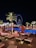 A nighttime scene of a luxurious outdoor pool area with orange umbrellas and loungers, illuminated by palm trees and accent lights. In the background, a large Ferris wheel glows with blue and white lights, enhancing the vibrant cityscape. The setting suggests a relaxing and upscale atmosphere.