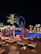 A nighttime scene of a luxurious outdoor pool area with orange umbrellas and loungers, illuminated by palm trees and accent lights. In the background, a large Ferris wheel glows with blue and white lights, enhancing the vibrant cityscape. The setting suggests a relaxing and upscale atmosphere.