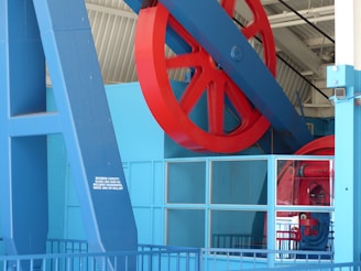 A large red wheel is mounted on a sturdy blue structure inside an industrial or mechanical setting. The architecture contains metal beams and grid-like windows, suggesting a factory or machinery room environment. A small sign indicates maximum capacity in both pounds and kilograms.