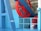 A large red wheel is mounted on a sturdy blue structure inside an industrial or mechanical setting. The architecture contains metal beams and grid-like windows, suggesting a factory or machinery room environment. A small sign indicates maximum capacity in both pounds and kilograms.