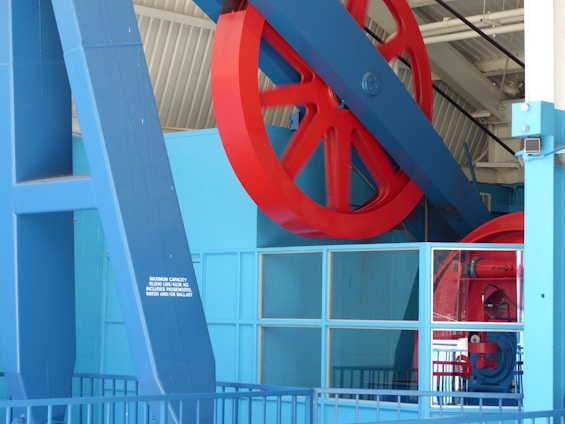 A large red wheel is mounted on a sturdy blue structure inside an industrial or mechanical setting. The architecture contains metal beams and grid-like windows, suggesting a factory or machinery room environment. A small sign indicates maximum capacity in both pounds and kilograms.