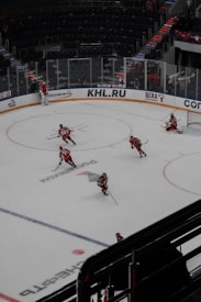Several hockey players wearing red and white uniforms are practicing on an indoor ice rink. The rink features markings and advertisements in various languages, and the surrounding stands are mostly empty. A goaltender is positioned near the net.