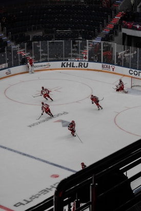Several hockey players wearing red and white uniforms are practicing on an indoor ice rink. The rink features markings and advertisements in various languages, and the surrounding stands are mostly empty. A goaltender is positioned near the net.