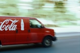 A red delivery van featuring a large beverage company logo and bottle icon is driving down a street. The motion blur effect indicates speed, with a slightly blurred background of trees and buildings visible.