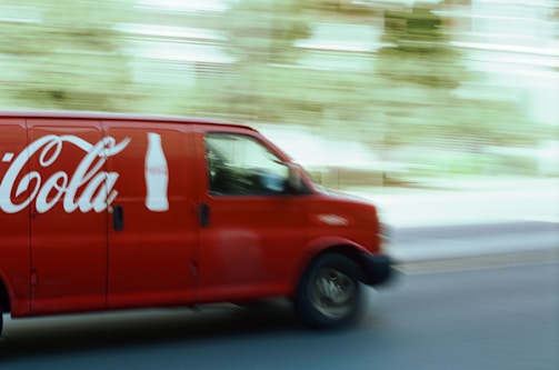 A red delivery van featuring a large beverage company logo and bottle icon is driving down a street. The motion blur effect indicates speed, with a slightly blurred background of trees and buildings visible.