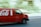 A red delivery van featuring a large beverage company logo and bottle icon is driving down a street. The motion blur effect indicates speed, with a slightly blurred background of trees and buildings visible.