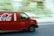 A red delivery van featuring a large beverage company logo and bottle icon is driving down a street. The motion blur effect indicates speed, with a slightly blurred background of trees and buildings visible.