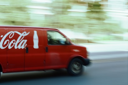 A red delivery van featuring a large beverage company logo and bottle icon is driving down a street. The motion blur effect indicates speed, with a slightly blurred background of trees and buildings visible.