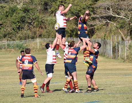 Rugby players are engaged in a line-out, with two teams wearing different colored jerseys. Players are lifted to catch the ball mid-air on a grassy field with trees and fencing in the background.