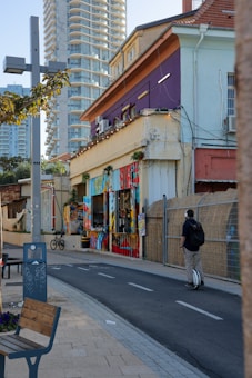 An urban street scene featuring a vibrant and colorful mural painted on the side of a building. The architecture includes both older low-rise structures and modern high-rise buildings in the background. A person is riding a unicycle on a path alongside the street. Bicycles and street furniture, such as a bench, are also visible. The setting appears lively and artistic.