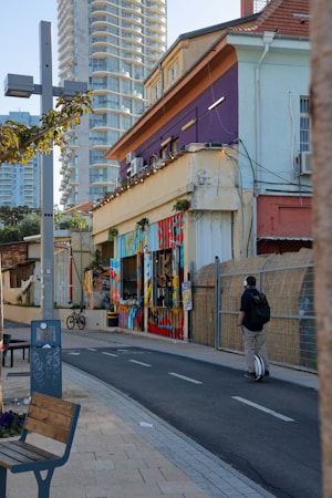 An urban street scene featuring a vibrant and colorful mural painted on the side of a building. The architecture includes both older low-rise structures and modern high-rise buildings in the background. A person is riding a unicycle on a path alongside the street. Bicycles and street furniture, such as a bench, are also visible. The setting appears lively and artistic.