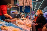 A bustling fish market with a vendor wearing a red sweater and a face mask, serving a customer who is also wearing a mask. The counter is filled with various types of seafood on ice. Additional customers can be seen in the background, and the market is well-lit with overhead lighting.