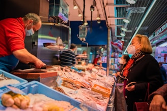 A bustling fish market with a vendor wearing a red sweater and a face mask, serving a customer who is also wearing a mask. The counter is filled with various types of seafood on ice. Additional customers can be seen in the background, and the market is well-lit with overhead lighting.