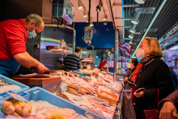 A bustling fish market with a vendor wearing a red sweater and a face mask, serving a customer who is also wearing a mask. The counter is filled with various types of seafood on ice. Additional customers can be seen in the background, and the market is well-lit with overhead lighting.