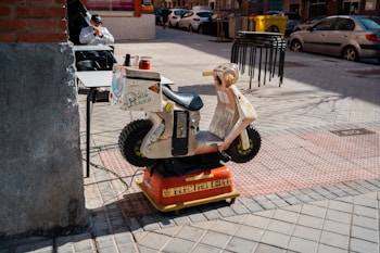 A small children's ride in the shape of a motorcycle is placed on a tiled sidewalk. The ride features bright colors, including red and yellow accents, and displays various stickers. Behind it, outdoor seating is visible with a man sitting at a table. There's a backdrop of parked cars and urban buildings.