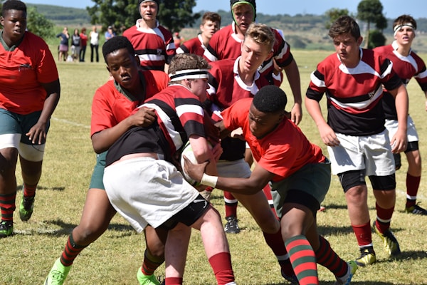 A group of young rugby players in action on a grassy field under a clear sky. Two players in red jerseys are attempting to tackle a player in a black, red, and white jersey. Other players in similar uniforms are watching or participating in the play.