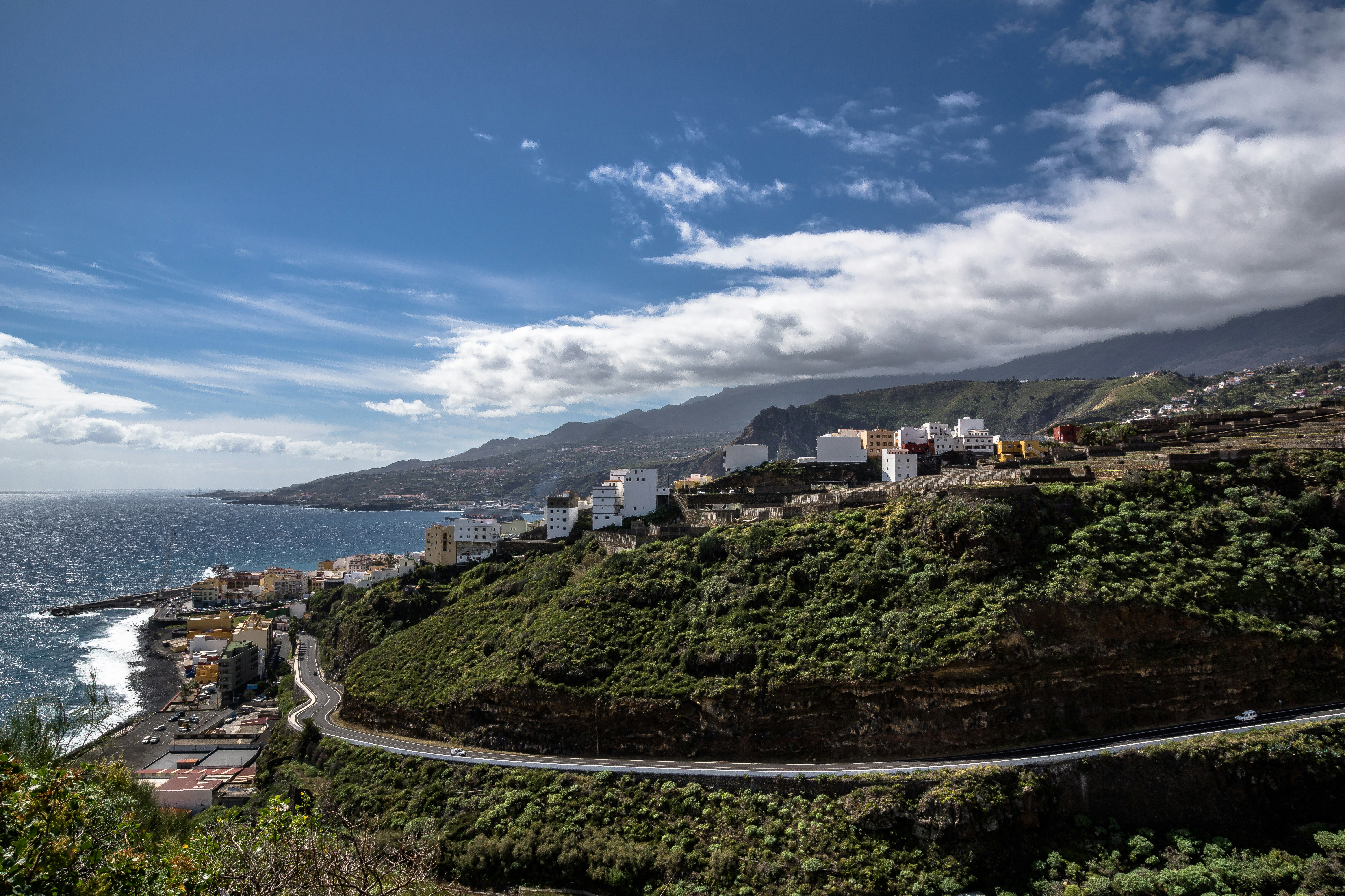 View of Santa Cruz and a coastal road of La Palma. Steep and wooded mountainsides (on which houses are partly built) flat down to the sea . The blue sky is partly covers by white clouds.