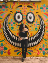 A smiling person wearing bold, geometric glasses standing in front of a city mural.