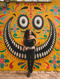 A smiling person wearing bold, geometric glasses standing in front of a city mural.