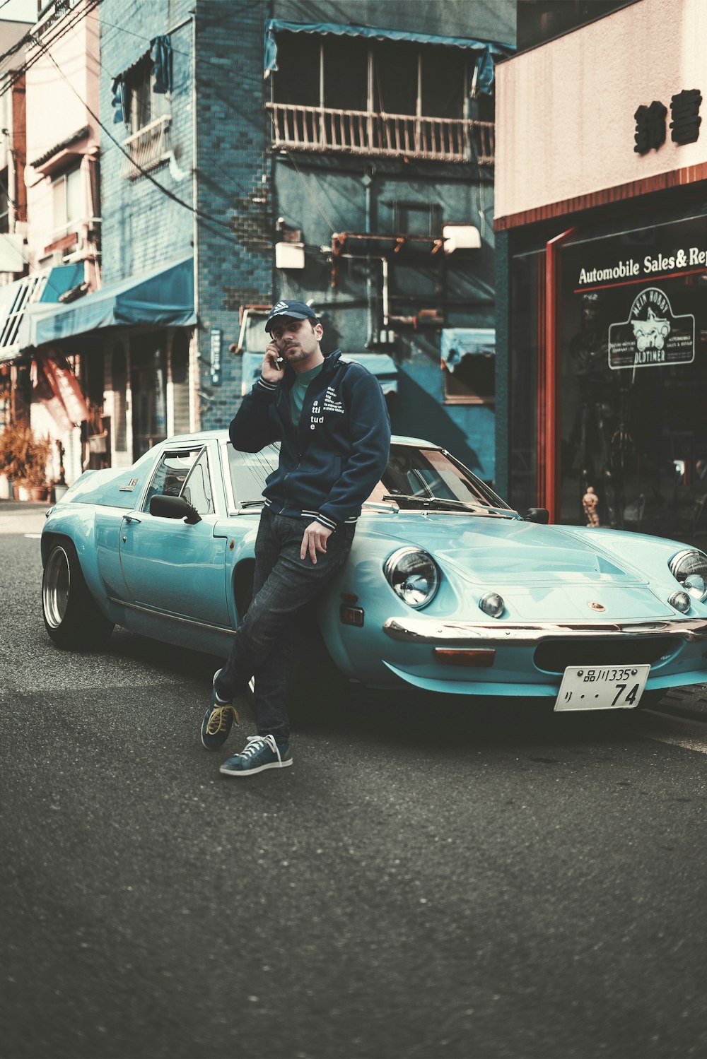 man in black jacket and black pants leaning on blue porsche 911 coupe during daytime