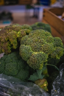Large bunches of fresh broccoli with dense, green florets sit prominently in focus. The background is slightly blurred, suggesting a market or grocery setting with wooden crates visible.