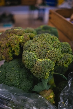 Large bunches of fresh broccoli with dense, green florets sit prominently in focus. The background is slightly blurred, suggesting a market or grocery setting with wooden crates visible.