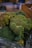 Large bunches of fresh broccoli with dense, green florets sit prominently in focus. The background is slightly blurred, suggesting a market or grocery setting with wooden crates visible.