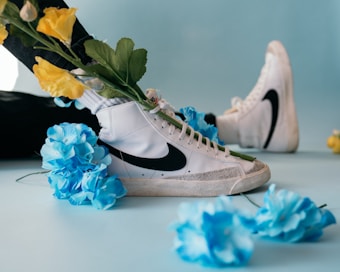 A pair of white high-top sneakers with black swoosh logos are adorned with vibrant blue and yellow flowers. The shoes are set against a light blue background, suggesting a playful and artistic composition.