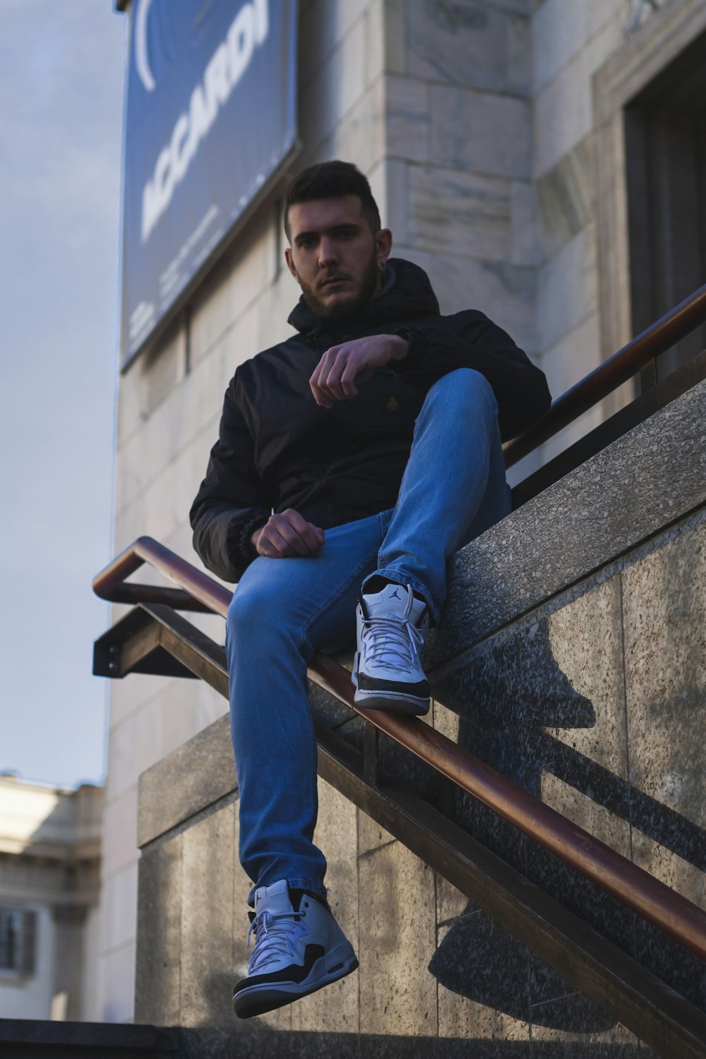 man in black jacket and blue denim jeans sitting on concrete stairs