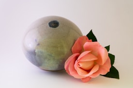 A smooth, round ceramic vase with a subtle gray and blue glaze sits on a simple white background. Beside it lies a delicate pink rose with lush green leaves, creating a harmonious and elegant composition.