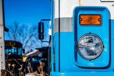 Close-up view of the front section of an old blue bus with visible headlight and side light. In the background, another bus is partially visible amidst a setting that includes dry vegetation and other vehicles. The sky is clear and blue.