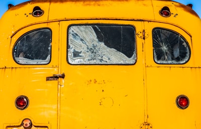 An old yellow vehicle with three cracked windows at the back. The paint is slightly worn, and the vehicle has vintage round red lights. Some rust is visible along the edges.